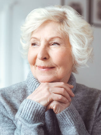 Older woman with white hair in a gray sweater, smiling and resting her hands under her chin, looking to the side.