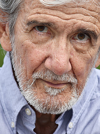 Older man with gray hair and beard wearing a light blue collared shirt, looking directly at the camera.