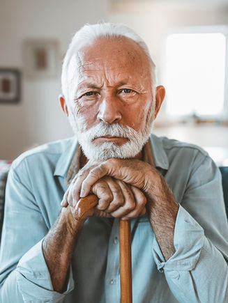 Older man with white hair and beard sits indoors, resting his hands and chin on a wooden cane, looking at the camera.