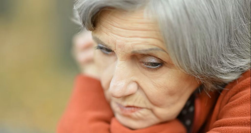 Elderly woman with gray hair and an orange sweater looks downward with a pensive expression.