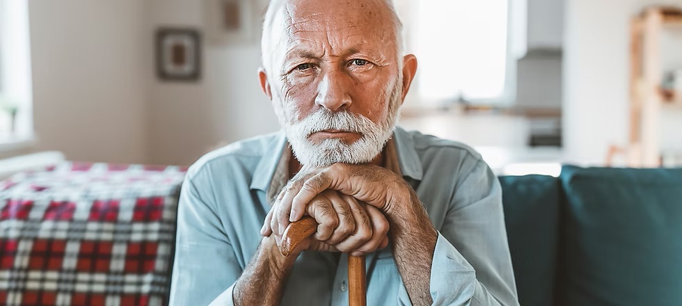 Elderly man with white hair and beard sits indoors, leaning on a wooden cane, looking toward the camera.