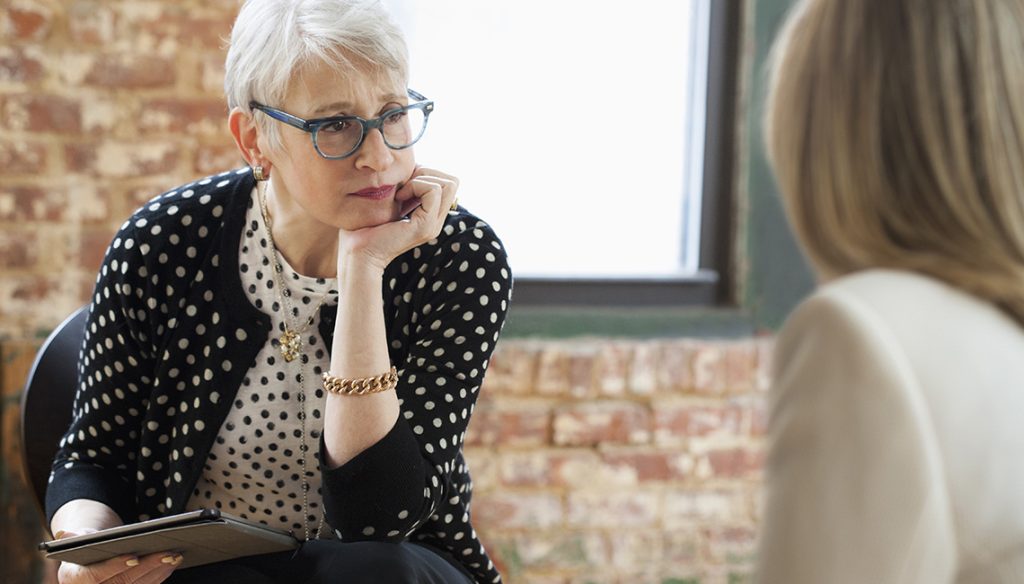 A woman with short gray hair and glasses listens attentively to another person during a conversation indoors.