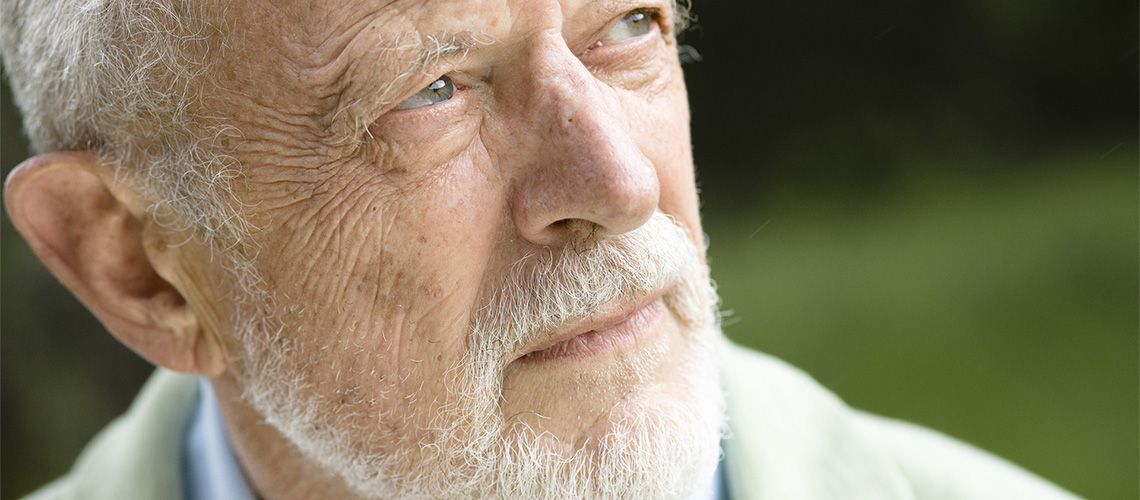 Close-up of an elderly man with a white beard and mustache looking thoughtfully into the distance outdoors.