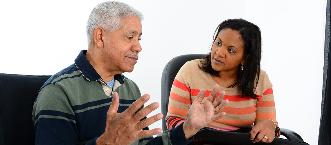 An older man speaks while a woman listens attentively, seated beside him in a conversation setting.