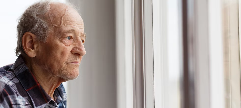 Elderly man in a plaid shirt looks out a window with a thoughtful expression.